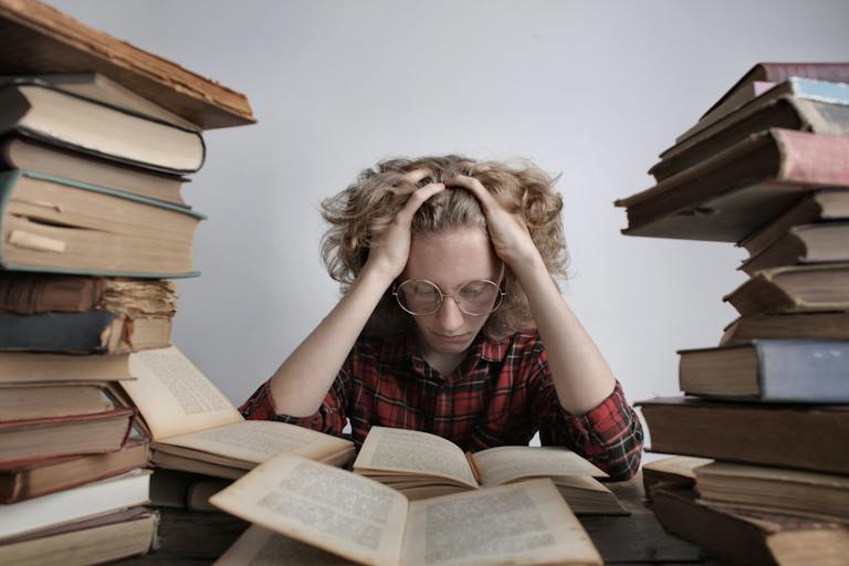 Tired teenage boy in casual clothes and eyeglasses sitting at desk with large stacks of books holding head and reading book while preparing for exam